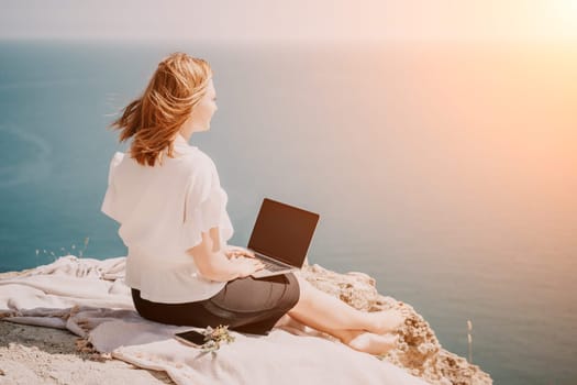 Successful business woman in yellow hat working on laptop by the sea. Pretty lady typing on computer at summer day outdoors. Freelance, travel and holidays concept.