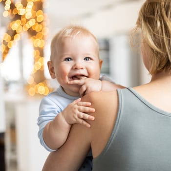 Blonde mother holding her adorable cheerful little infant baby boy. Cute infant child looking in camera and smiling at home