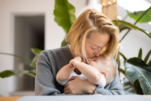 Portrait of young mother cuddling and kissing her adorable little baby boy while sitting at the table at home. Sensory stimulation for baby development