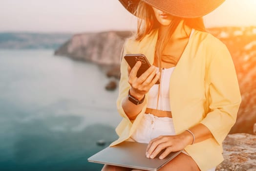 Successful business woman in yellow hat working on laptop by the sea. Pretty lady typing on computer at summer day outdoors. Freelance, travel and holidays concept.