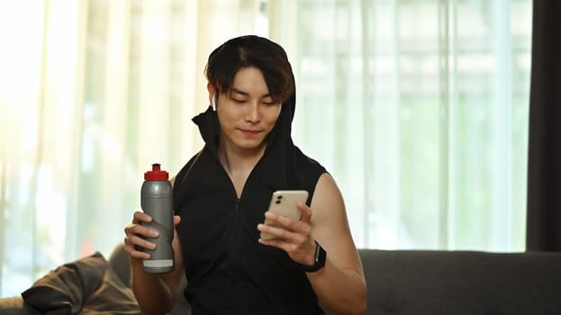 Young asian male athlete checking training program on smartphone application and drinking water while having break.