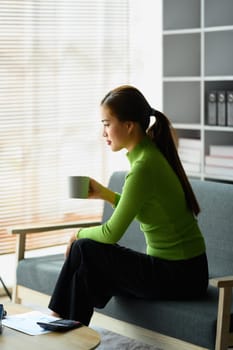 Satisfied millennial woman is sitting on couch at home with cup of coffee. People, relaxing and leisure concept.