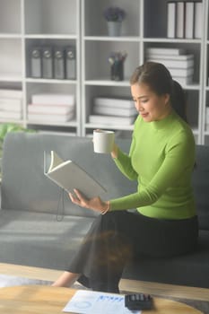 Calm young woman drinking hot tea and reading book on comfortable sofa. People, leisure and lifestyle concept.