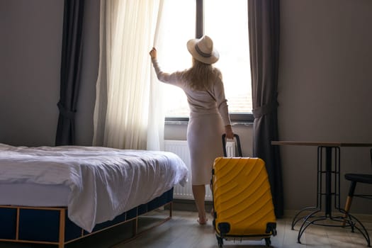 Portrait of tourist woman standing nearly window, looking to beautiful view with her luggage in hotel bedroom after check-in.