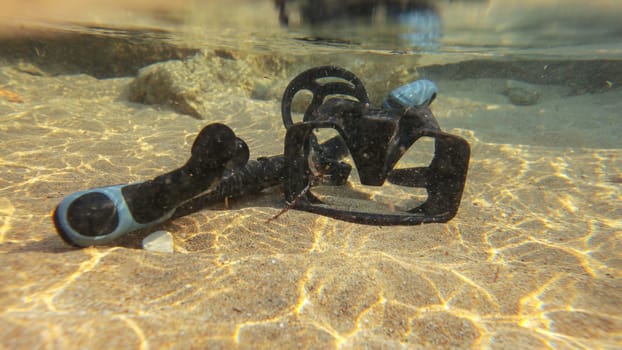 Underwater photo, black snorkel and diving mask on sand in shallow water, sun shining.