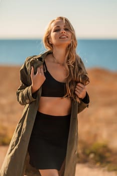 Portrait blonde sea cape. A calm young blonde in an unbuttoned khaki raincoat stands on the seashore, under the raincoat there is a black skirt and top.