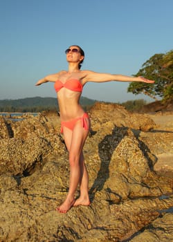 Young slim woman in bikini swim wear and sunglasses, standing on the rocks near beach, arms spread, facing to sun, as if gathering energy.