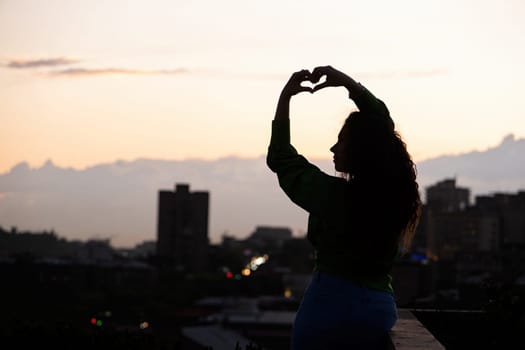 silhouette of beautiful woman on rooftop at urban sunset, making heart shape with hands