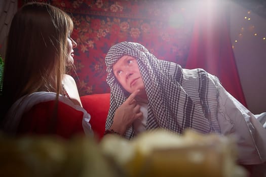 Portrait of young arabian muslim couple in traditional clothes in cozy red room. Fhoto shoot in easten style with male and female model like in harem with a sultan and an odalisque. Partial focus