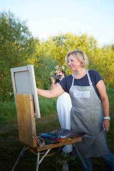 Adult female artist painting picture near water of river or lake in nature. Girl in white sundress and flower wreath for holiday of Ivan Kupala in nature at sunset near artist