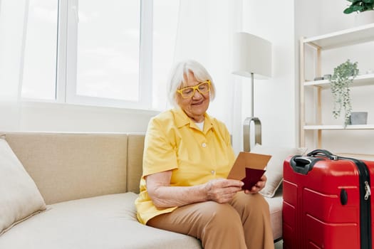 Happy senior woman with passport and travel ticket packed a red suitcase, vacation and health care. Smiling old woman joyfully sitting on the sofa before the trip raised her hands up in joy. High quality photo