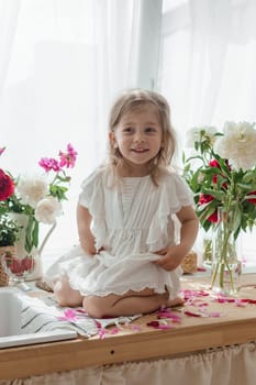 A little blonde girl on a kitchen countertop decorated with peonies. Spring atmosphere