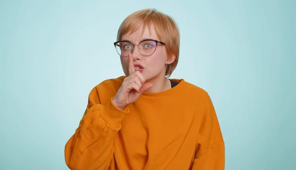 Shh be quiet please. Young woman presses index finger to lips makes silence gesture sign do not tells secret, stop gossip. Pretty short haired blonde girl isolated alone on blue studio background