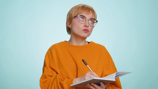 Thoughtful journalist woman making notes, writing down thoughts with pen into notepad notebook diary, to do list, good idea. Pretty short haired blonde girl isolated alone on blue studio background