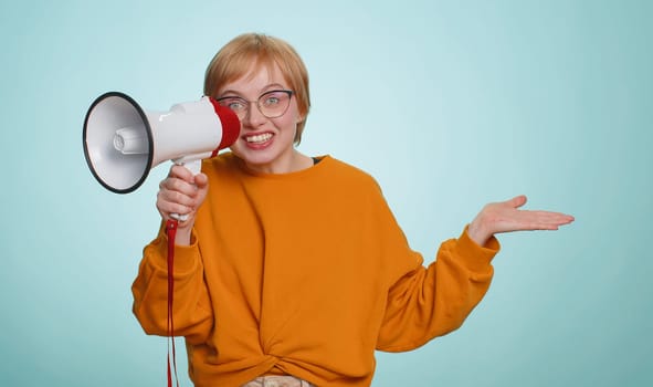 Woman talking with megaphone, proclaiming news, loudly announcing advertisement, warning using loudspeaker to shout speech, pointing empty place. Copy-space. Pretty blonde girl on blue wall background