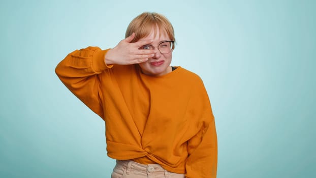 Nosy curious young woman in glasses closing eyes with hand and spying through fingers, hiding and peeping, binocular gesture exploring way seeking something in distance. Blonde girl on blue background