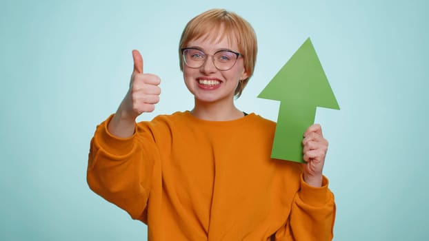 Young woman winner holding arrow sign pointing up, career growth and money exchange increase. Blonde girl showing up indicating successful development progress isolated on blue studio background