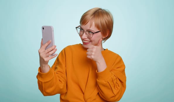 Woman blogger in glasses, taking selfie vlog on smartphone, communicating video call online with family or friends, live translation stream. Pretty blonde girl isolated on blue background, indoors