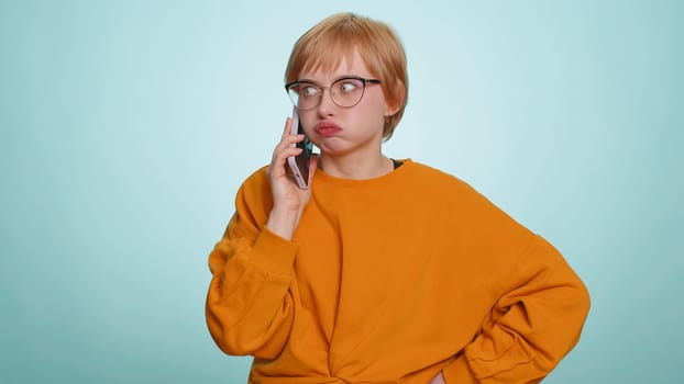 Tired sleepy young woman in glasses talking on mobile phone with friend making online conversation. Disinterested short haired girl having annoyed boring talk on smartphone isolated on blue background