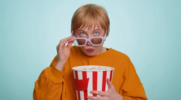 Excited young woman in 3D glasses eating popcorn and watching interesting tv serial, sport game, film, online social media movie content. Blonde short haired girl isolated on blue studio background