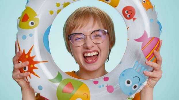 Close-up portrait of funny young tourist woman looking out of rubber swimming ring hole smiling enjoying summer vacation time. Travel, recreation, sea journey. Blonde girl isolated on blue background