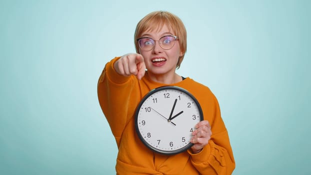 It is your time. Young woman in glasses showing time on wall office clock, ok, thumb up, approve, start, pointing finger at camera. Blonde short haired girl isolated on blue studio background, indoors
