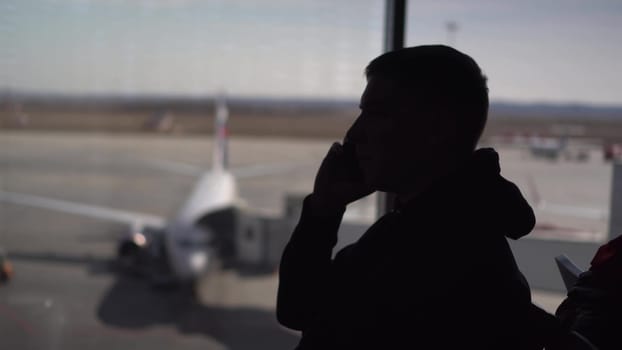 A young man in a sweatshirt is sitting at the airport and talking on the phone. Silhouette of a man on the background of the planes outside the window. 4k