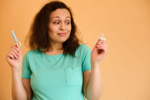 Multi ethnic young pretty brunette, charming pregnant woman in blue turquoise t-shirt, holding a baby pacifier and nail scissors, isolated on orange background. Pregnancy. Childbirth. Maternity leave