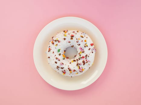Tasty doughnutdonut, top view. Sweet food leftlovers. Homemade circle donut with white icing and choclate rainbow sprinkles on a trendy pink background and white plate situated in the center of image.