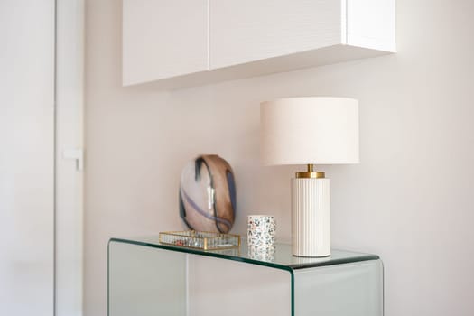 Close-up of a glass console with accessories and a lamp with a hanging white cabinet in the hallway or living room. Interior detail concept in modern style.