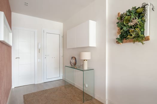 Simple but cozy entrance hall in light colors with an intercom, a glass console and a hanging cabinet with a stylish herbarium on the wall. The concept of simple concise interior design.