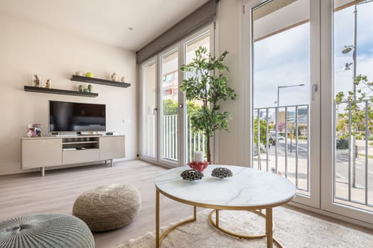 Comfortable living room with large windows and natural light with large TV, table and ottomans. The concept of a functional simple and stylish interior.