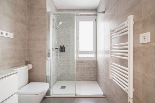 Comfortable bathroom with a toilet bowl and a shower cabin with tiles in beige tones and a window for natural light. The concept of a bathroom in a hotel or apartment after renovation.
