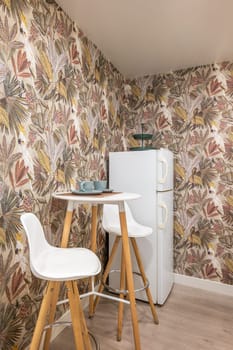 Compact high small round table with cups and two bar stools in the kitchen on the backdrop of boho style wallpaper and a refrigerator. Concept of a small design space.