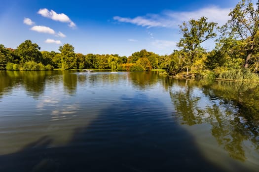 Beautiful sunny panorama of big lake with small fountains at center and green trees and bushes around in city South park 