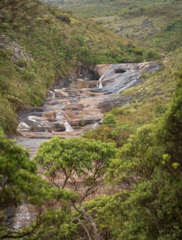 Unrecognizable people walk through the stone basin of the river, exploring different colorful pools of the Enchanted Valley in Caparao, Brazil. Solid rock eroded by years of flowing water, surrounded by lush forest, perfect for social media.