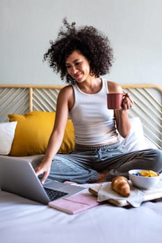 Happy young multiracial latina woman sitting on bed typing on laptop while having morning coffee. Vertical image. Lifestyle concept.