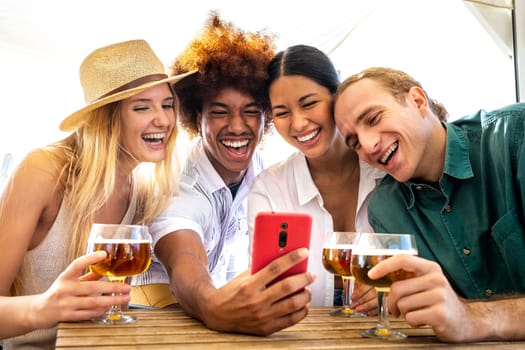 Group of happy multiracial friends looking at mobile phone in a beach bar while drinking beer. Friends on a video call using phone. Taking selfie. Technology and leisure activity.