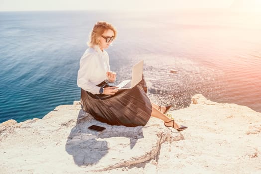 Digital nomad, Business woman working on laptop by the sea. Pretty lady typing on computer by the sea at sunset, makes a business transaction online from a distance. Freelance, remote work on vacation