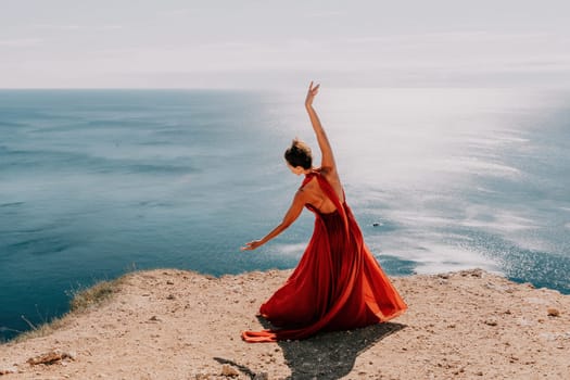 Side view a Young beautiful sensual woman in a red long dress posing on a rock high above the sea during sunrise. Girl on the nature on blue sky background. Fashion photo.