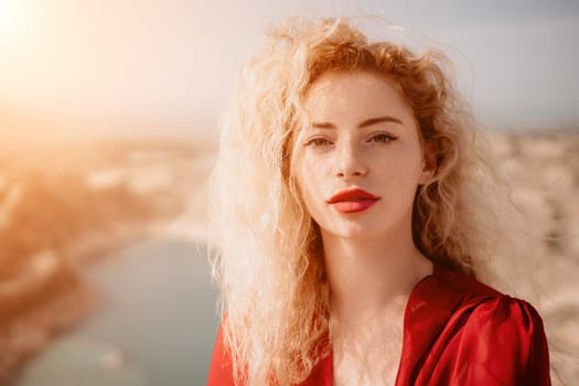 Close up shot of beautiful young caucasian woman with curly blond hair and freckles looking at camera and smiling. Cute woman portrait in a pink long dress posing on a volcanic rock high above the sea