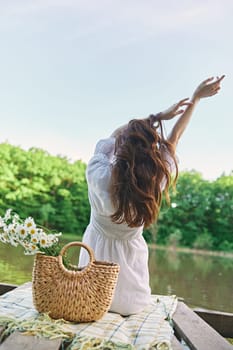 a woman sits on a blanket near the lake with her hands raised above her head and looks at the view. High quality photo