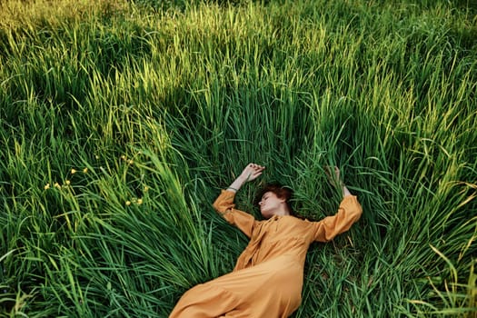 a sweet, calm woman in an orange dress lies in a green field with her arms outstretched, enjoying the silence and peace. Horizontal photo taken from above. High quality photo