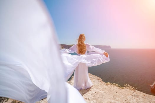 woman sea white dress. Blonde with long hair on a sunny seashore in a white flowing dress, rear view, silk fabric waving in the wind. Against the backdrop of the blue sky and mountains on the seashore