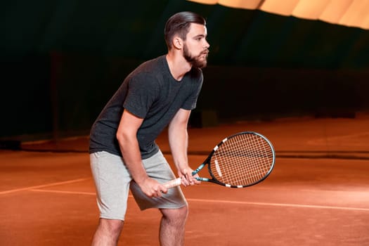 Handsome young man in t-shirt holding tennis racket and looking concentrated while standing on tennis court