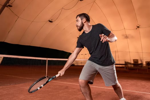 Handsome young man in t-shirt holding tennis racket and looking concentrated while standing on tennis court