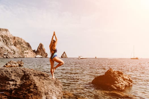 Yoga on the beach. A happy woman meditating in a yoga pose on the beach, surrounded by the ocean and rock mountains, promoting a healthy lifestyle outdoors in nature, and inspiring fitness concept