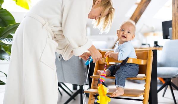 Happy infant sitting and playing with his toy in traditional scandinavian designer wooden high chair in modern bright atic home suppervised by his mother