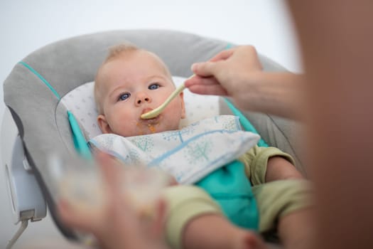 Mother spoon feeding her baby boy child in baby chair with fruit puree. Baby solid food introduction concept