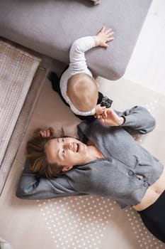 Happy family moments. Mother lying comfortably on children's mat playing with her baby boy watching and suppervising his first steps. Positive human emotions, feelings, joy